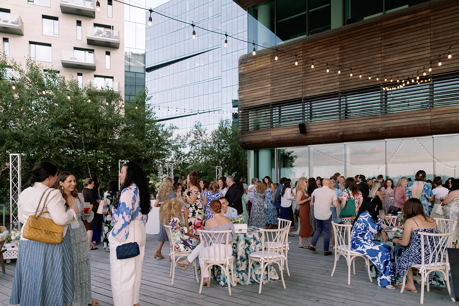 A large group of people dressed in summer attire gather on a modern outdoor terrace, socializing and sitting at tables under string lights, with tall buildings and greenery in the background.