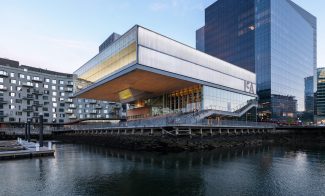 A modern waterfront building with large glass windows and an overhanging upper floor, set beside a dock and surrounded by contemporary office and apartment buildings. The scene is reflected in the calm water.