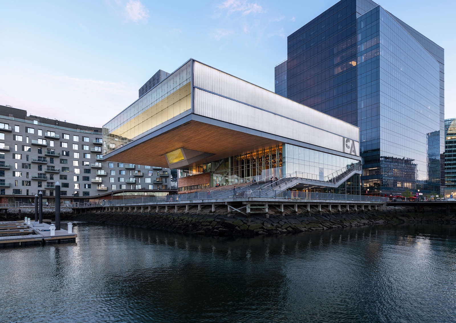 A modern waterfront building with large glass windows and an overhanging upper floor, set beside a dock and surrounded by contemporary office and apartment buildings. The scene is reflected in the calm water.