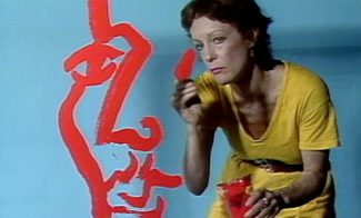 A woman in a yellow dress paints a large red abstract face on a light blue wall. She is holding a paintbrush in one hand and a red paint container in the other, looking intently at her artwork.