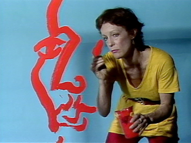 A woman in a yellow dress paints a large red abstract face on a light blue wall. She is holding a paintbrush in one hand and a red paint container in the other, looking intently at her artwork.