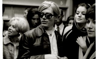 A black-and-white photo shows a man in large, dark sunglasses and a leather jacket surrounded by a group of stylishly dressed women and men at a crowded indoor gathering.
