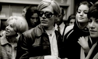 A man with light hair and dark sunglasses wears a leather jacket, standing among a group of people who appear to be socializing at the crowded ICA 90 YEARS indoor event.
