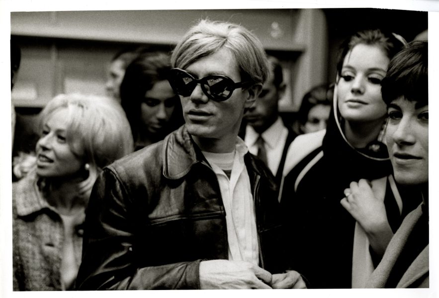 A black-and-white photo shows a man in large, dark sunglasses and a leather jacket surrounded by a group of stylishly dressed women and men at a crowded indoor gathering.