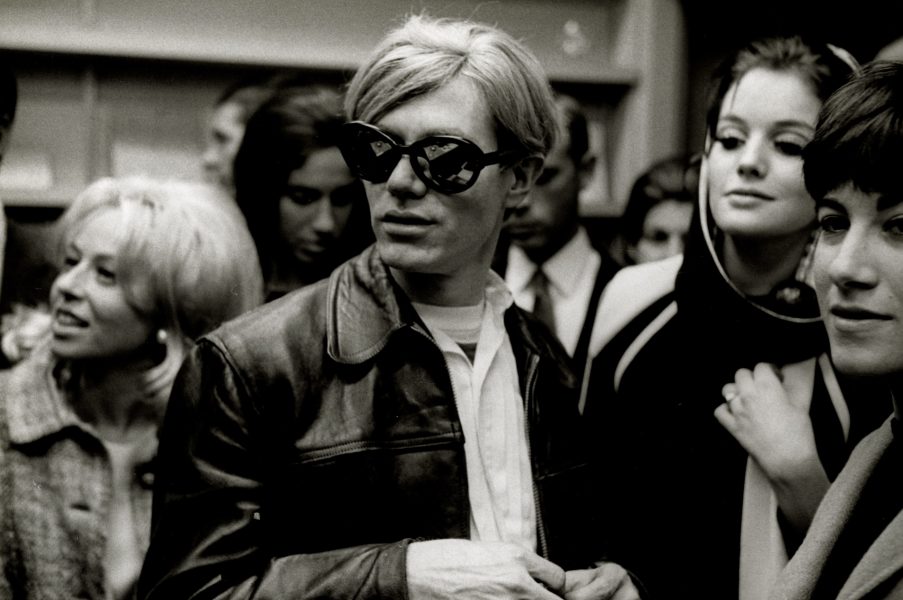 A man with light hair and dark sunglasses wears a leather jacket, standing among a group of people who appear to be socializing at the crowded ICA 90 YEARS indoor event.