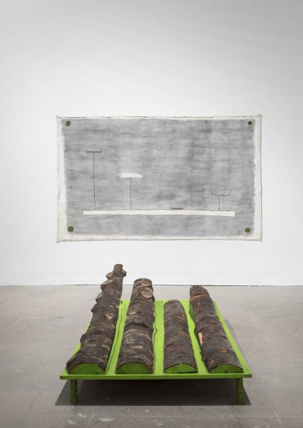 A minimalist art installation features three logs placed on a bright green platform. Behind it, a grayscale abstract drawing with lines and simple shapes is displayed on the wall. The setting is a clean, white-walled gallery.