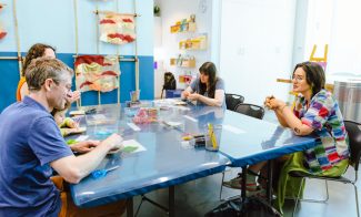Four adults sit around a blue table in the ICA Art Lab classroom, working on crafts with supplies as part of the Perfect Memory workshop. Artworks and shelves with materials are visible in the background.