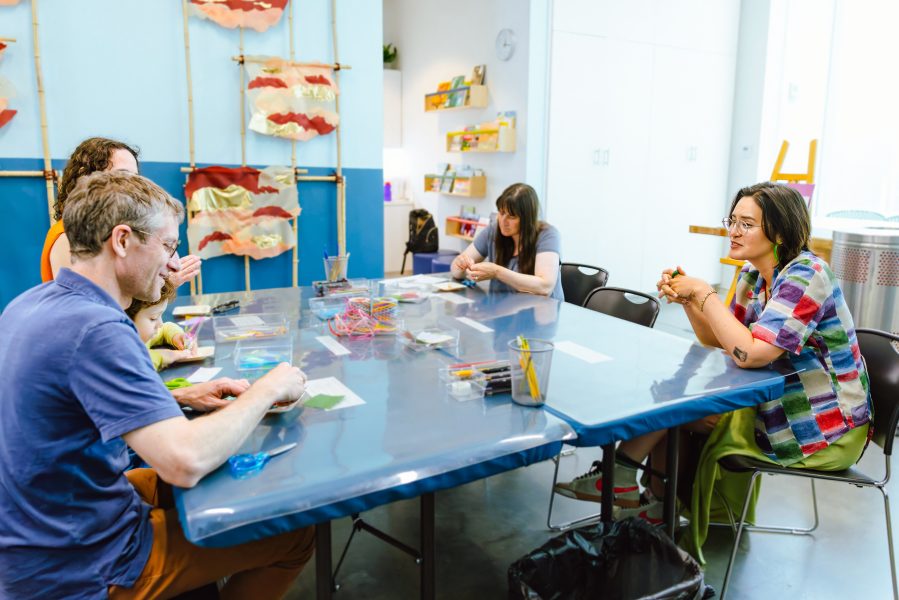 Four adults sit around a blue table in the ICA Art Lab classroom, working on crafts with supplies as part of the Perfect Memory workshop. Artworks and shelves with materials are visible in the background.