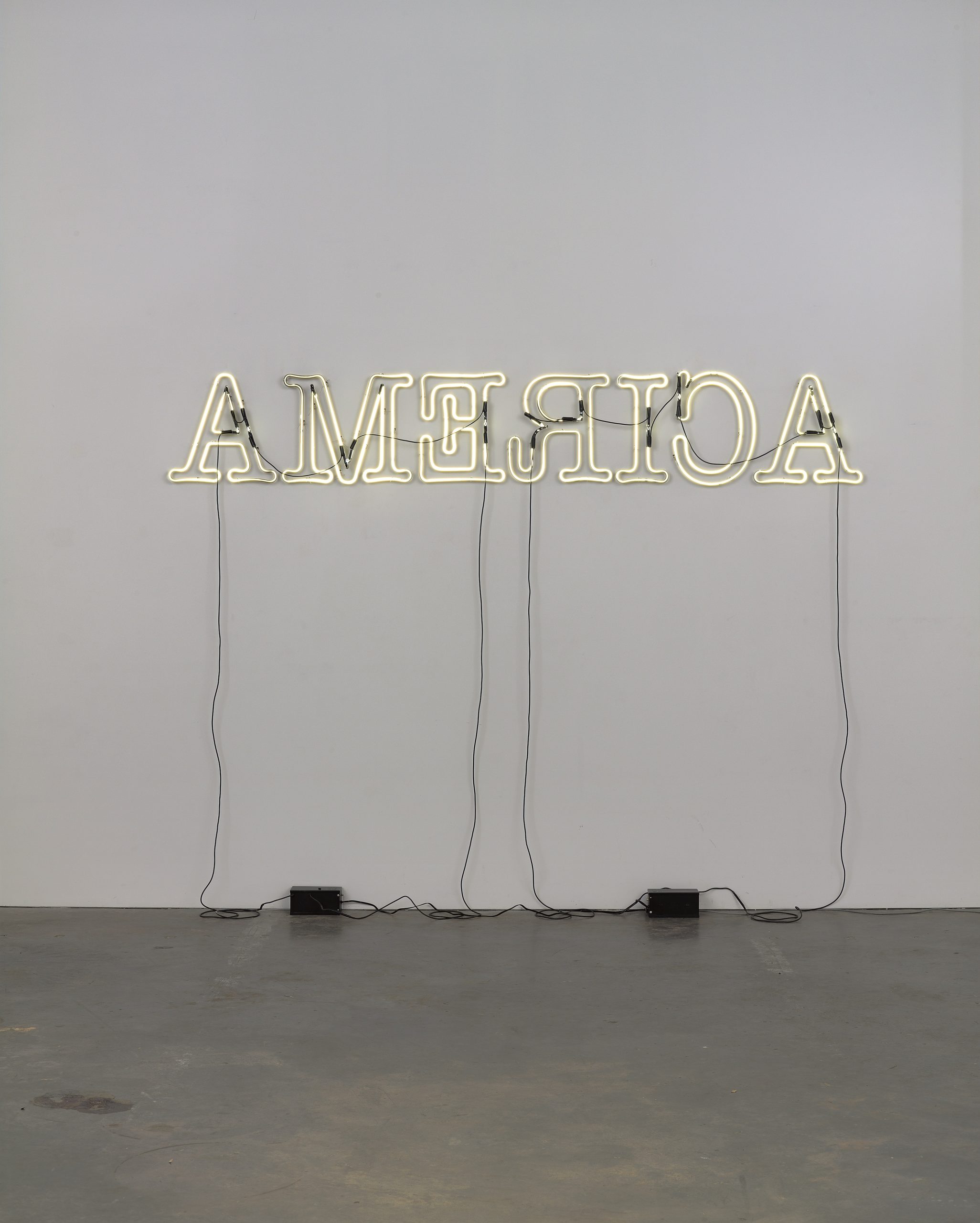 A neon sign spells AMERICA in all capital letters, but the text is displayed backwards against a plain white wall with cords hanging down to three black boxes on the floor.