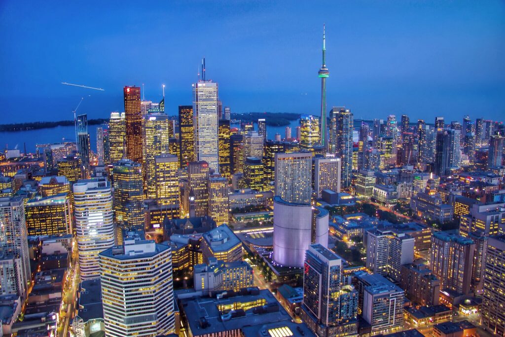 A vibrant city skyline at dusk with tall buildings illuminated by lights, including a prominent tower with a green-lit spire, under a clear blue sky.