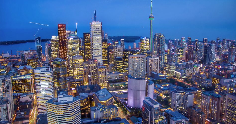 A vibrant city skyline at dusk with tall buildings illuminated by lights, including a prominent tower with a green-lit spire, under a clear blue sky.