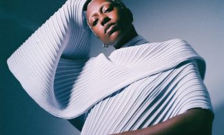 A person poses in a sculptural, futuristic, ribbed white garment with dramatic folds and textures, one arm bent over their head. The lighting casts soft shadows, creating an artistic and modern atmosphere.