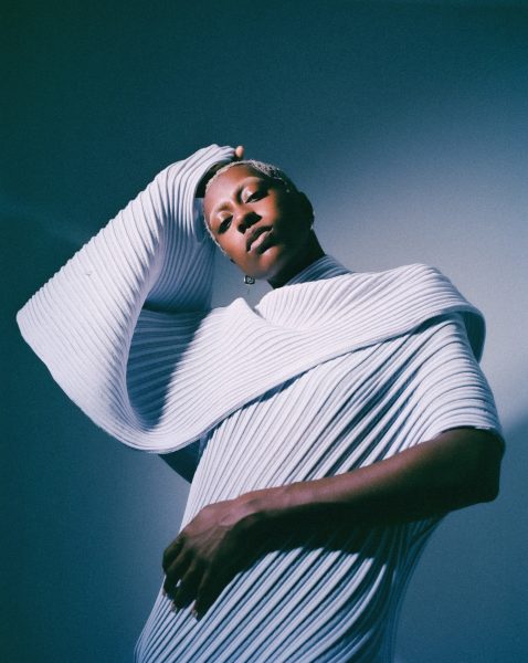 A person poses in a sculptural, futuristic, ribbed white garment with dramatic folds and textures, one arm bent over their head. The lighting casts soft shadows, creating an artistic and modern atmosphere.