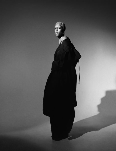 A person with short hair stands confidently in a dramatic black outfit, looking slightly to the side. The black-and-white image features strong lighting, casting a distinct shadow on the studio floor and background.