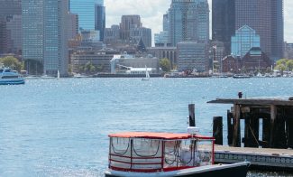 A small boat with a red roof sails near a dock on a sunny day, with a city skyline featuring tall glass buildings in the background across the water.