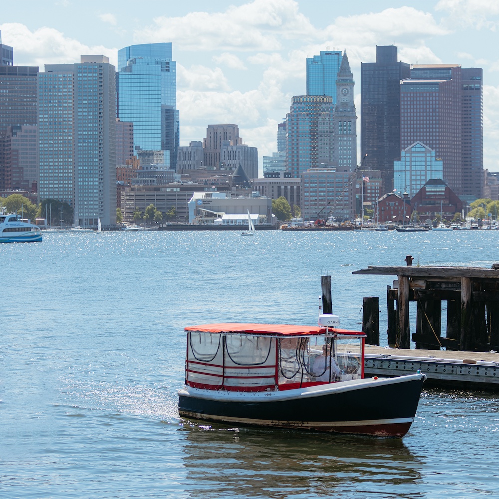 A small boat with a red roof sails near a dock on a sunny day, with a city skyline featuring tall glass buildings in the background across the water.