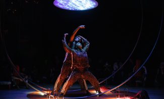 Two dancers in textured costumes pose dramatically under a glowing, circular light, framed by a large metal hoop. Highlighted against a dark background with a few small lanterns on the floor nearby, their sweat variant adds a palpable sense of intensity and realism to the scene.