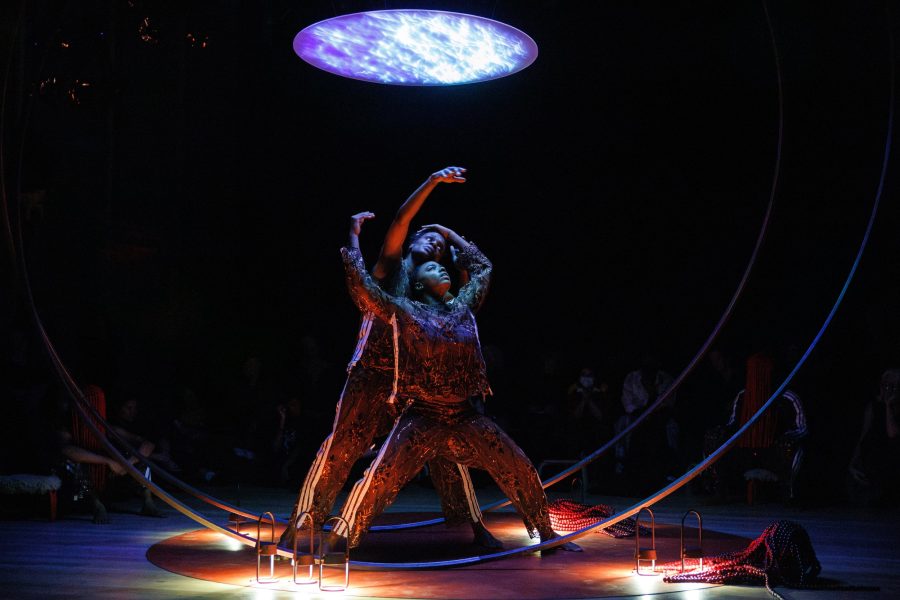 Two dancers in textured costumes pose dramatically under a glowing, circular light, framed by a large metal hoop. Highlighted against a dark background with a few small lanterns on the floor nearby, their sweat variant adds a palpable sense of intensity and realism to the scene.