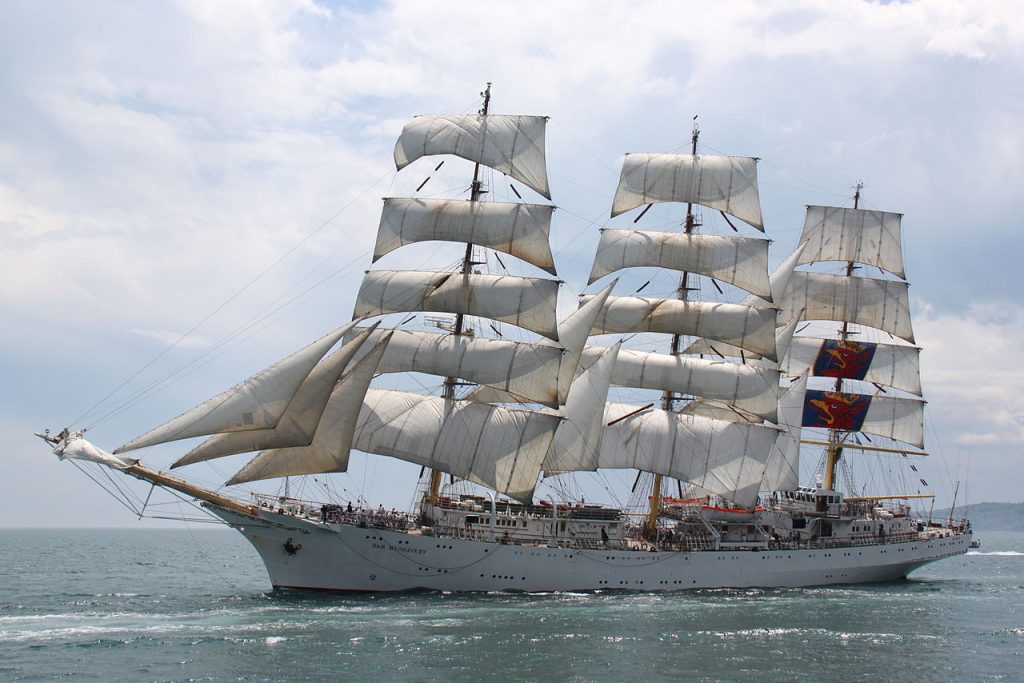 A large white Tall Ships sailing with multiple tall masts and many white sails fully unfurled, sails on the open sea under a partly cloudy sky. Crowds of people are visible on deck.