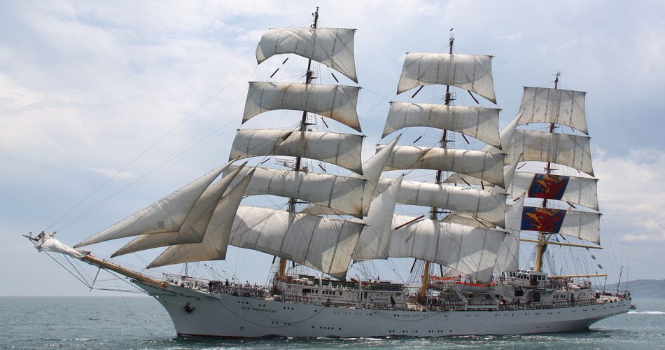 A large white Tall Ships sailing with multiple tall masts and many white sails fully unfurled, sails on the open sea under a partly cloudy sky. Crowds of people are visible on deck.