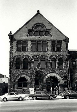 Black and white photo of a historic stone building with arched windows and three parked cars in front. Several people stand near the entrance, where a sign reads Museum of Contemporary Art, reminiscent of the classic architecture once associated with the Institute of Contemporary Art Boston (ICA Boston).