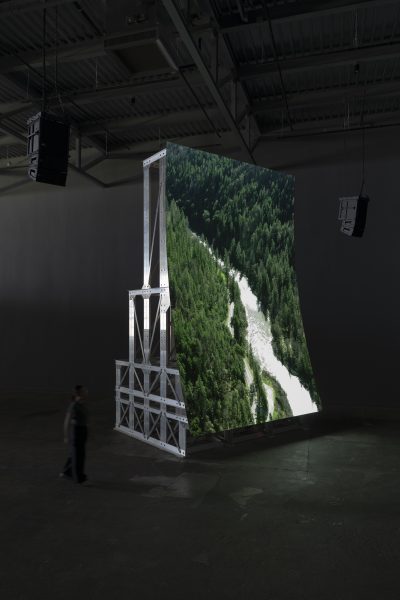 A large, freestanding screen displays an aerial view of a river running through a dense green forest. The screen is supported by a metal frame in a dark, industrial indoor space, with a person walking nearby.