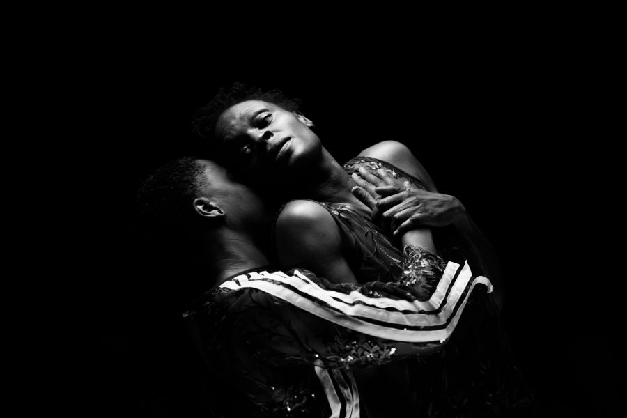 Two people embrace in dramatic lighting against a black background. One tilts their head back, eyes closed, while the other leans in closely, creating an intimate and emotional scene accented by a subtle sweat variant that heightens the raw intensity of their connection.