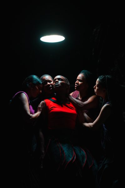Five people, in a sweat variant of emotional intensity, stand closely together under a single overhead light in a dark setting. One person in the center, dressed in red and glistening slightly as if from exertion or nerves, looks upward while the others touch or support them emotionally.