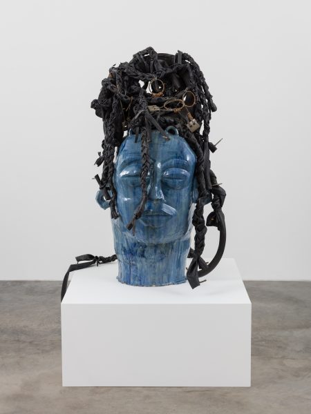 A large blue ceramic sculpture of a human head with eyes closed, adorned with black braided and twisted materials, sits on a white pedestal in a gallery space.