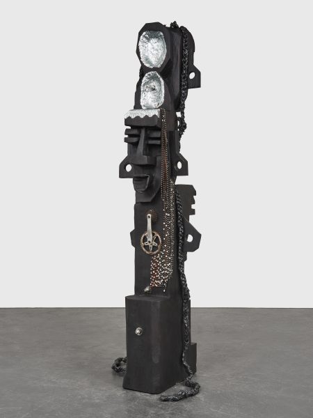 A tall, abstract black sculpture with geometric shapes, metallic chains, gears, and two silver foil surfaces near the top, standing on a gray floor against a plain white background.
