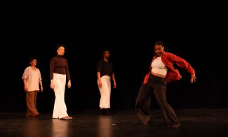 Four people stand on stage; three watch as one in front, wearing a red shirt, dramatically poses mid-dance against a dark background.