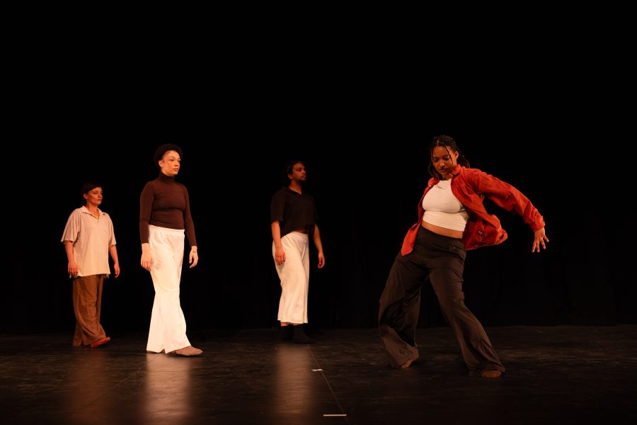 Four people stand on stage; three watch as one in front, wearing a red shirt, dramatically poses mid-dance against a dark background.