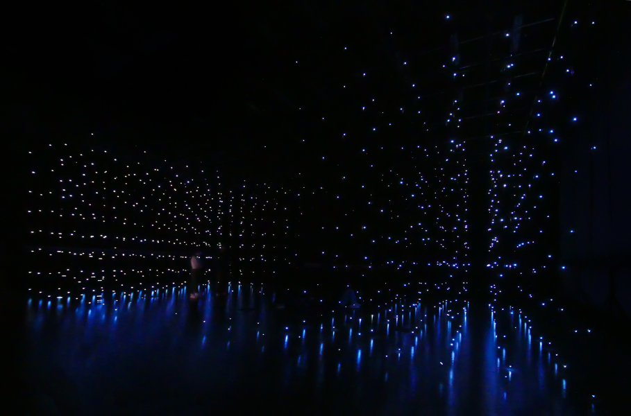 Dark room filled with countless small blue lights, some hanging from the ceiling and others reflecting off the shiny floor, creating a mesmerizing, starry, immersive space—nowiswhenweare the stars.