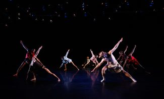 Eight dancers on a dark stage, each extending one arm and one leg outward in a diagonal pose, with dramatic lighting highlighting their movements. Small lights hang overhead.