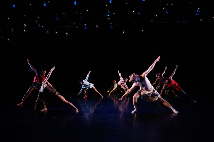 Eight dancers on a dark stage, each extending one arm and one leg outward in a diagonal pose, with dramatic lighting highlighting their movements. Small lights hang overhead.