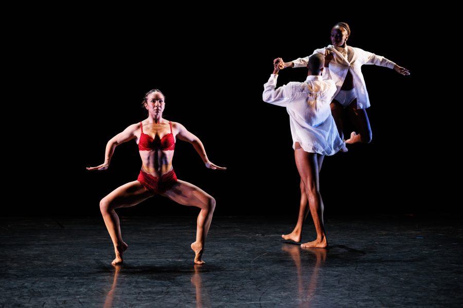 Three dancers perform on stage. One in a red outfit poses low with bent knees, while two others in white shirts interact mid-movement behind her. The background is dark, highlighting their forms.