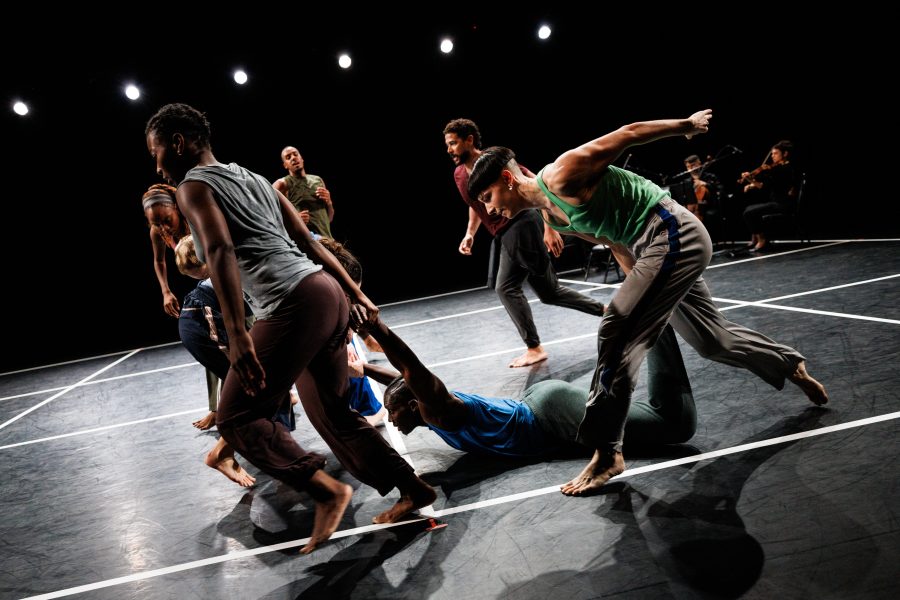 A group of barefoot dancers move energetically across a dark, stage-lit floor with white lines, while musicians play in the background.
