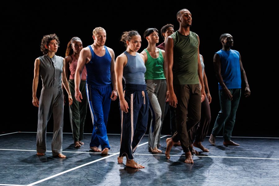 A diverse group of dancers in casual attire stand barefoot on a stage, facing forward, with serious expressions, under bright stage lighting against a black background.