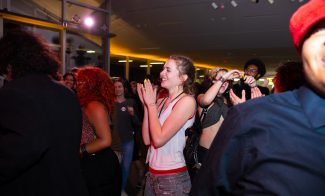 A group of people stand close together at an indoor event; a woman in the center is clapping and smiling while others around her appear to be enjoying themselves as well.