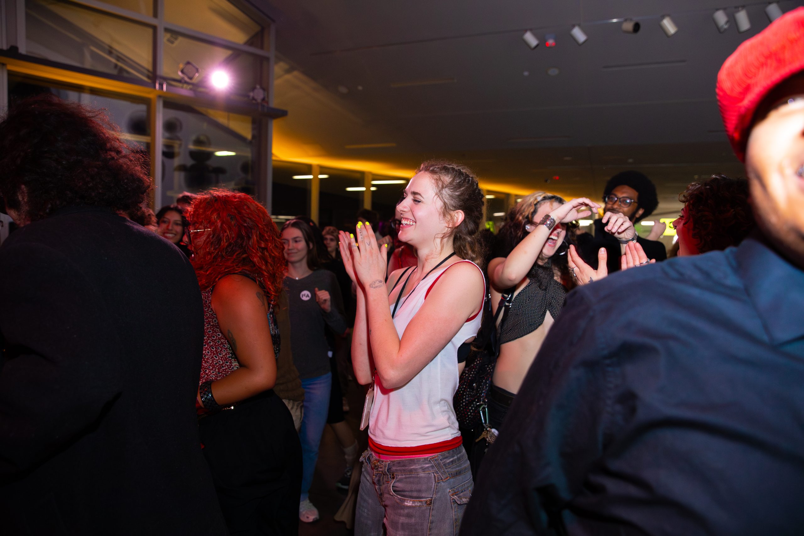 A group of people stand close together at an indoor event; a woman in the center is clapping and smiling while others around her appear to be enjoying themselves as well.