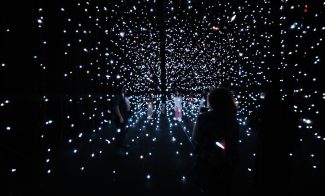 People walk through a dark room filled with suspended points of white light, creating a starry, immersive installation art experience—nowiswhenweare the stars.