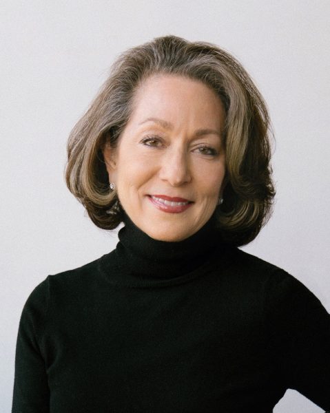 A woman with shoulder-length brown hair and a slight smile wears a black turtleneck sweater and stud earrings, standing against a plain light background.