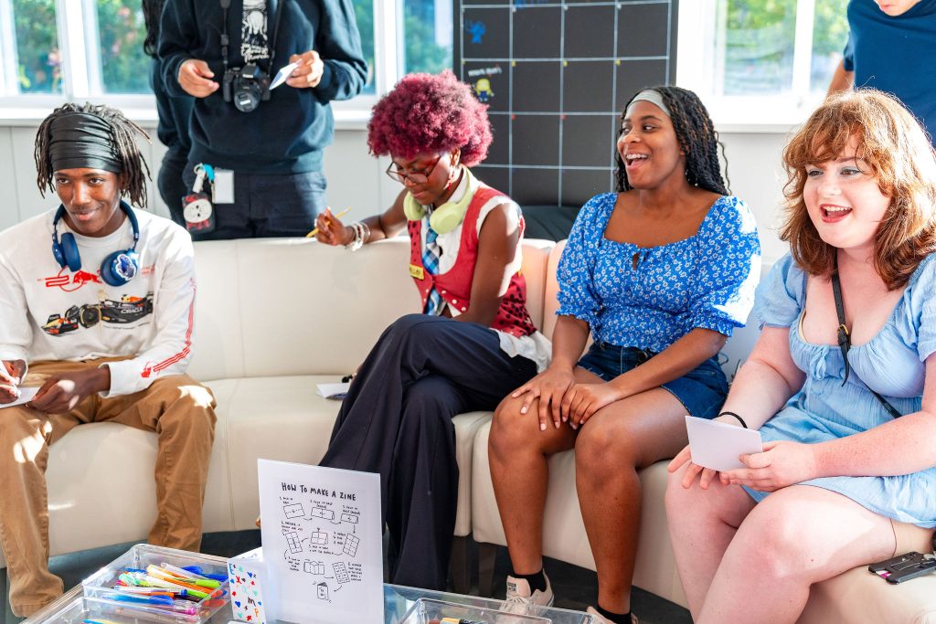 Four teens sit smiling on a white couch, holding papers and markers. A sign on the table says How to Make a Zine. Bright sunlight fills the room, creating a cheerful atmosphere.