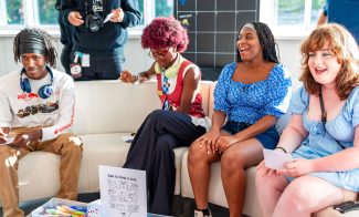 Four teens sit smiling on a white couch, holding papers and markers. A sign on the table says How to Make a Zine. Bright sunlight fills the room, creating a cheerful atmosphere.