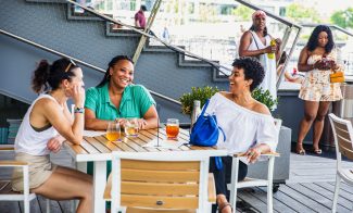 Three women sit at an outdoor table, smiling and talking over drinks, while two women stand in the background. The scene is lively and relaxed on a sunny day.
