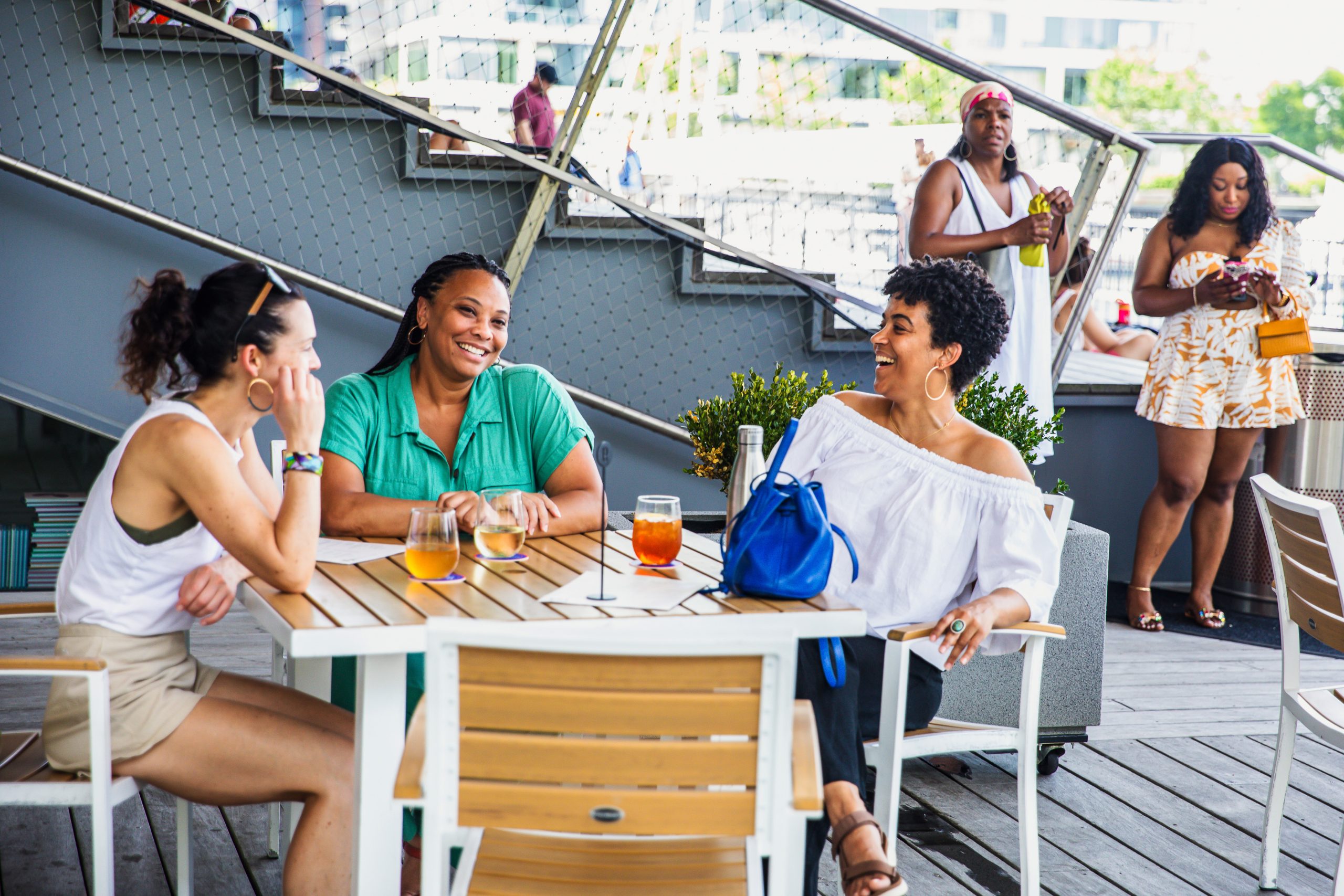 Three women sit at an outdoor table, smiling and talking over drinks, while two women stand in the background. The scene is lively and relaxed on a sunny day.