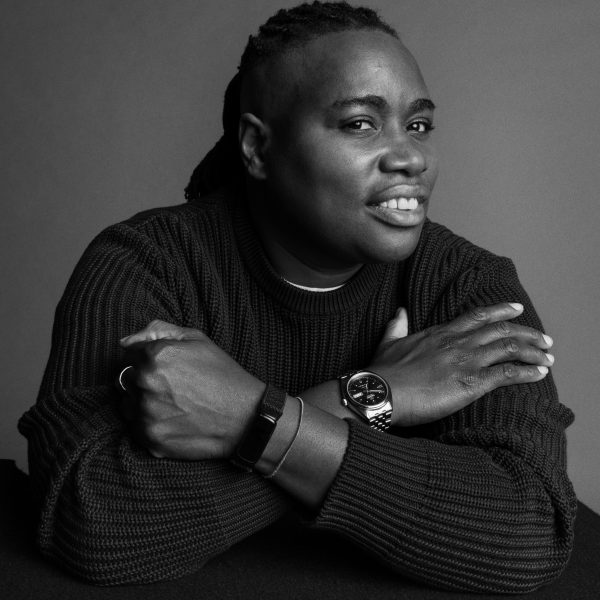 A person with short, styled hair and a ribbed sweater smiles at the camera, resting their crossed arms on a table. They wear a watch and bracelet, and the photo is in black and white.