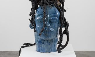 A large blue ceramic head sculpture with closed eyes and braided black fabric intertwined with objects arranged as hair, displayed on a white pedestal in a gallery setting.