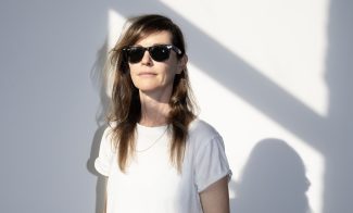A woman wearing sunglasses, a white t-shirt, and beige pants stands against a white wall with sunlight casting diagonal shadows. She has one hand in her pocket and a relaxed expression.
