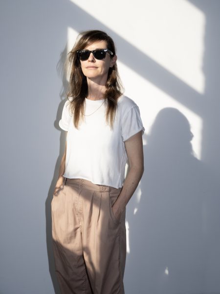 A woman wearing sunglasses, a white t-shirt, and beige pants stands against a white wall with sunlight casting diagonal shadows. She has one hand in her pocket and a relaxed expression.